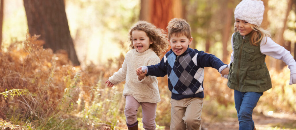 Three young happy children holding hands running through woods
