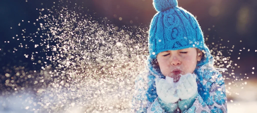 Young child in blue bobble hat blowing snow from gloves