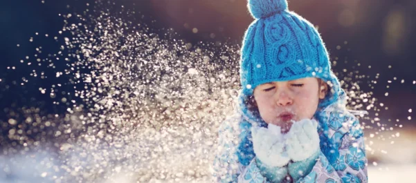 Young child in blue bobble hat blowing snow from gloves
