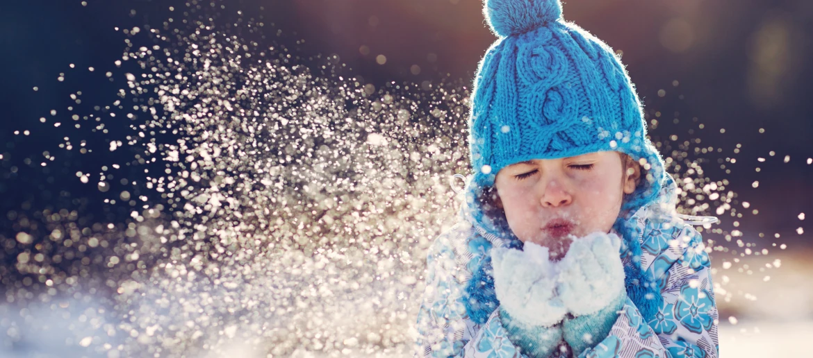 Young child in blue bobble hat blowing snow from gloves